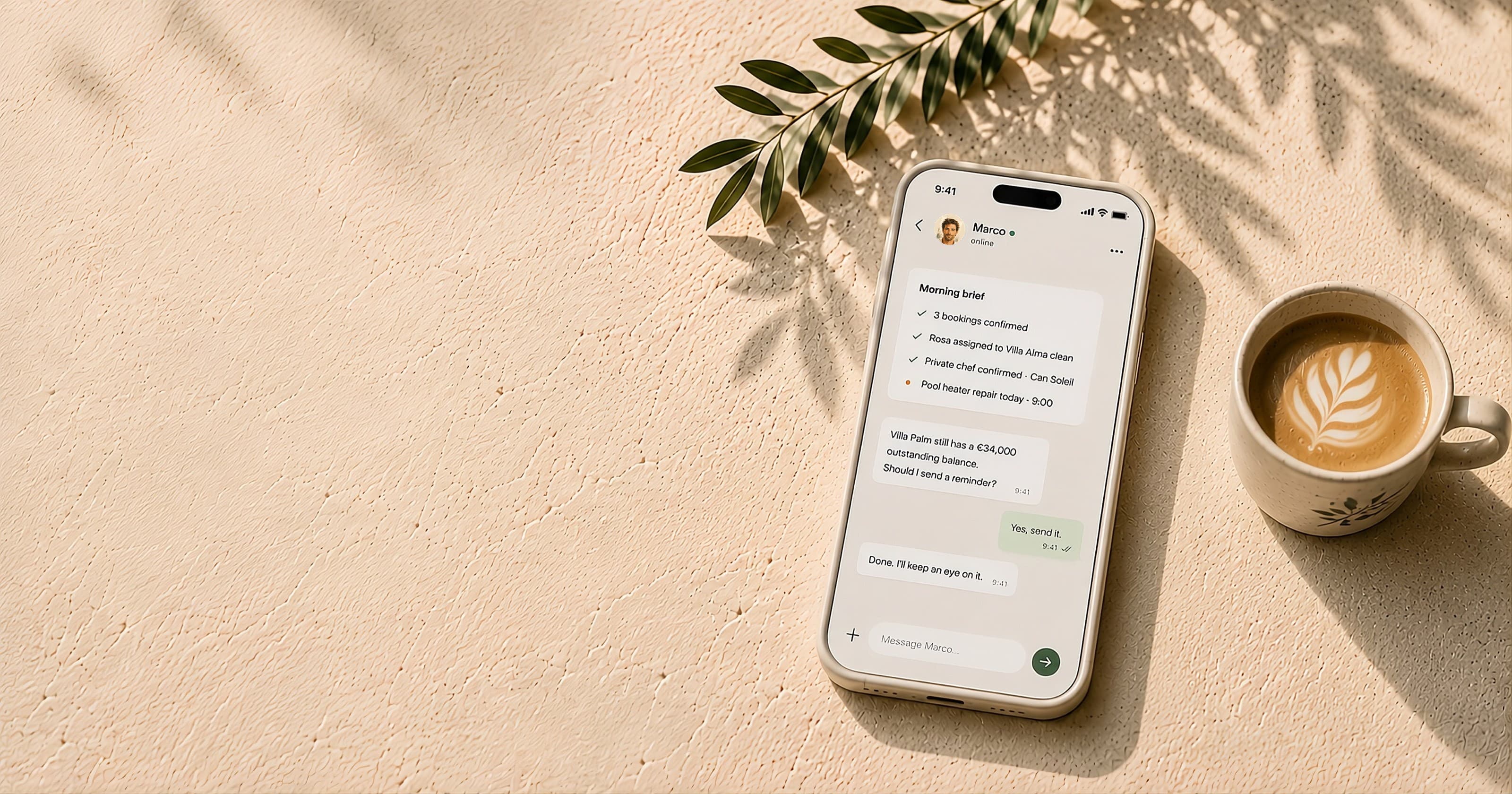 A phone on a sunlit Mediterranean stone table showing Marco's morning brief in WhatsApp, beside an olive branch and a glass of water